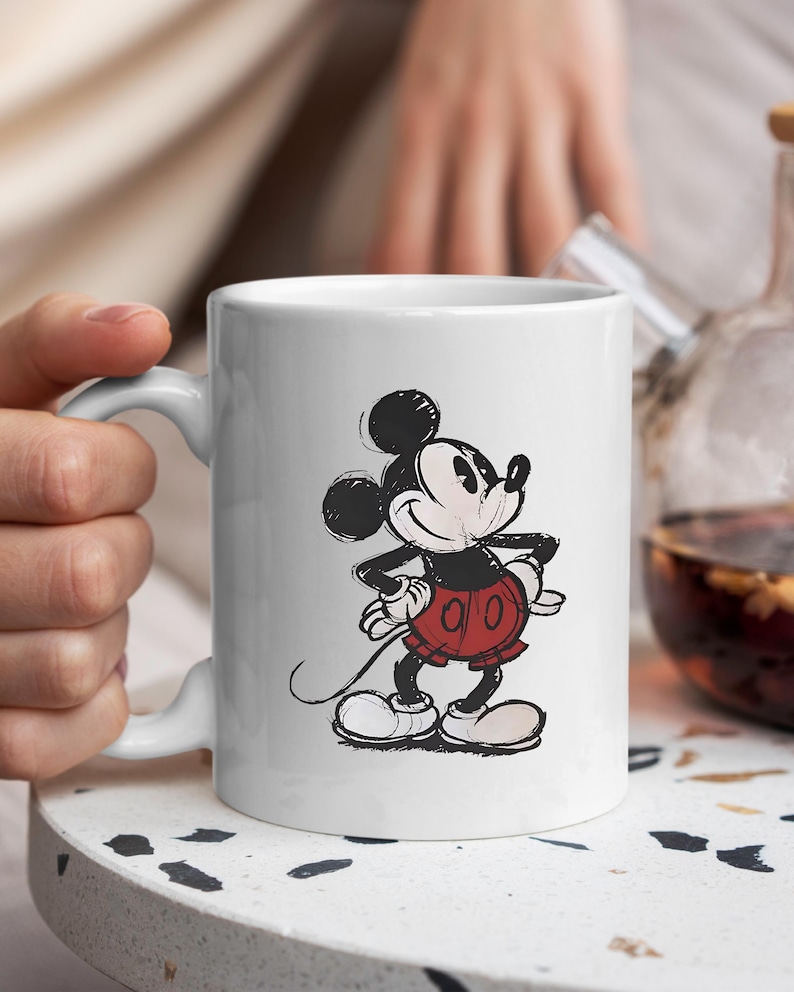 May include: White ceramic mug with a sketched illustration of Mickey Mouse. The cartoon character is shown in his classic outfit: red shorts, white gloves, and large white shoes. The mug has a curved handle and is held by a person.