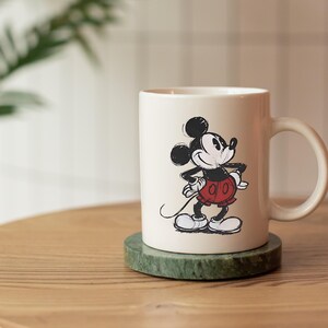 May include: Cream ceramic mug with a black and white Mickey Mouse illustration. The cartoon mouse is in his iconic pose, wearing red shorts and white gloves. The mug rests on a green marble coaster, a classic design.