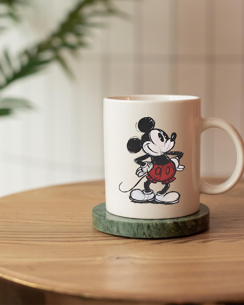 May include: Cream ceramic mug with a black and white Mickey Mouse illustration. The cartoon mouse is in his iconic pose, wearing red shorts and white gloves. The mug rests on a green marble coaster, a classic design.