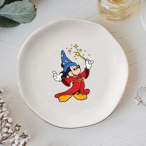 May include: A white ceramic plate with a gold rim featuring a cartoon image of Mickey Mouse dressed as a wizard. Mickey is wearing a blue hat, red robe, and yellow shoes, holding a wand with stars.