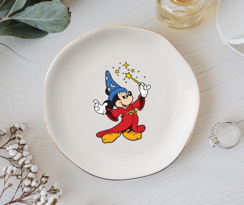 May include: A white ceramic plate with a gold rim featuring a cartoon image of Mickey Mouse dressed as a wizard. Mickey is wearing a blue hat, red robe, and yellow shoes, holding a wand with stars.
