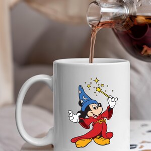 May include: White ceramic mug with a cartoon image of Mickey Mouse as a wizard. Mickey is in a blue hat with a moon and stars, a red robe, and yellow shoes. He holds a wand with stars. The mug is being filled with a dark liquid.