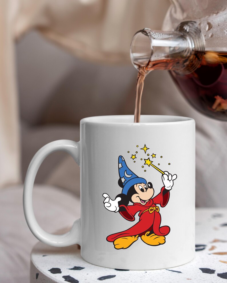 May include: White ceramic mug with a cartoon image of Mickey Mouse as a wizard. Mickey is in a blue hat with a moon and stars, a red robe, and yellow shoes. He holds a wand with stars. The mug is being filled with a dark liquid.
