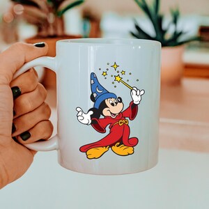 May include: White ceramic mug with a colourful illustration of Mickey Mouse as a wizard. Mickey wears a blue hat with stars, a red robe, and yellow shoes. He holds a wand with stars. The mug has a white handle. The design is from a popular cartoon.