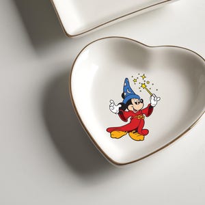 May include: A heart-shaped, white ceramic trinket dish with a gold rim. The dish features a colorful illustration of Mickey Mouse dressed as a wizard, holding a wand with stars. A second, rectangular dish is visible in the background.