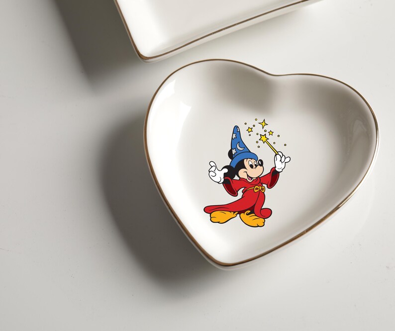 May include: A heart-shaped, white ceramic trinket dish with a gold rim. The dish features a colorful illustration of Mickey Mouse dressed as a wizard, holding a wand with stars. A second, rectangular dish is visible in the background.