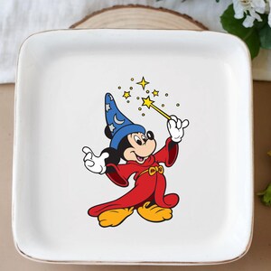 May include: White square ceramic plate featuring a cartoon image of Mickey Mouse dressed as a wizard. Mickey is wearing a blue hat with stars, a red robe, and yellow shoes, holding a wand with yellow stars.