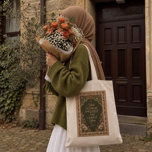 May include: A beige canvas tote bag featuring a rectangular graphic with an ornate floral border and the text "And he found you LOST And Guided You" in a decorative font. The bag is carried by someone wearing an olive green sweater and white skirt.