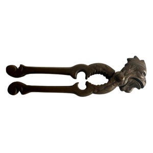 May include: A dark bronze-toned metal nutcracker featuring a sculpted rooster head at the hinge. The tool has two long, curved handles with decorative scrollwork ends and a serrated interior grip area designed for cracking shells.