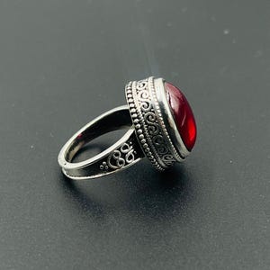 Op de afbeelding: Een zilveren ring met een ovale, dieprode edelsteen. De ring heeft sierlijke, wervelende patronen rond de edelsteen en op de band. De ring staat tegen een donkere achtergrond, waardoor de details worden benadrukt.