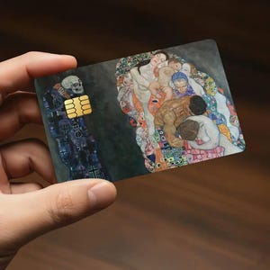 May include: A credit card featuring a detail from a Gustav Klimt painting. The card displays a skull on the left and a group of figures on the right, with gold-colored EMV chip. The artwork is set against a dark background.