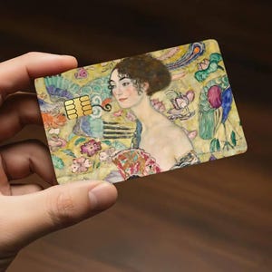 May include: A credit card featuring a painting of a woman with a floral and peacock feather background. The card has a gold chip and is held in a hand.
