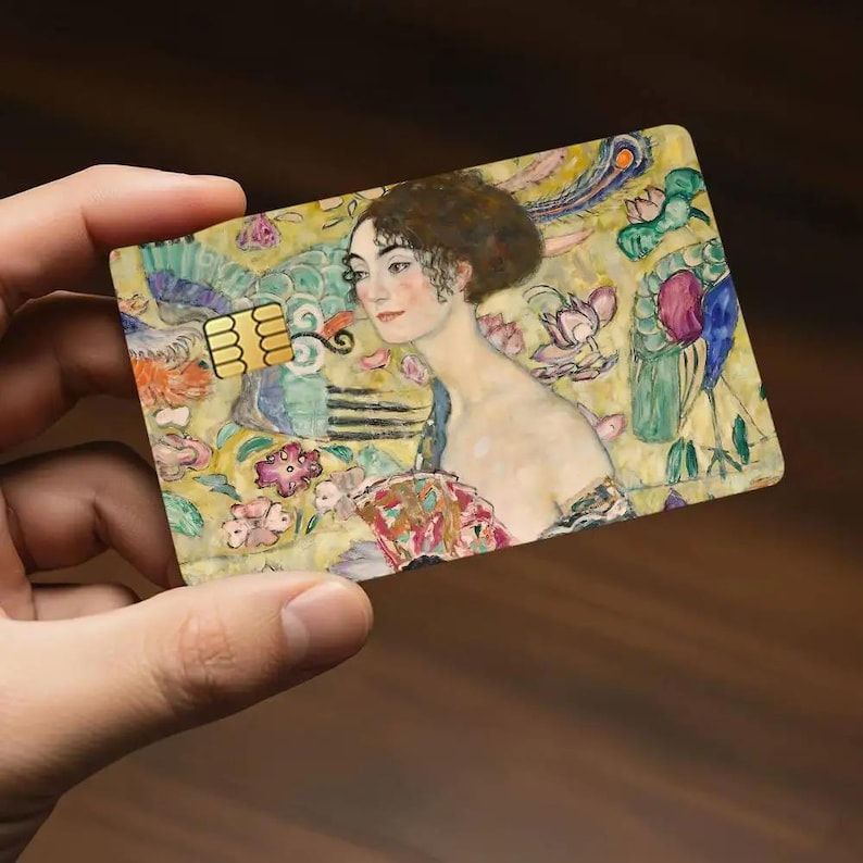 May include: A credit card featuring a painting of a woman with a floral and peacock feather background. The card has a gold chip and is held in a hand.