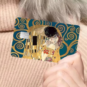 May include: A credit card featuring Gustav Klimt's "The Kiss" artwork. The card has a teal background with gold swirls and the iconic image of the embracing couple. The card also has a white chip.
