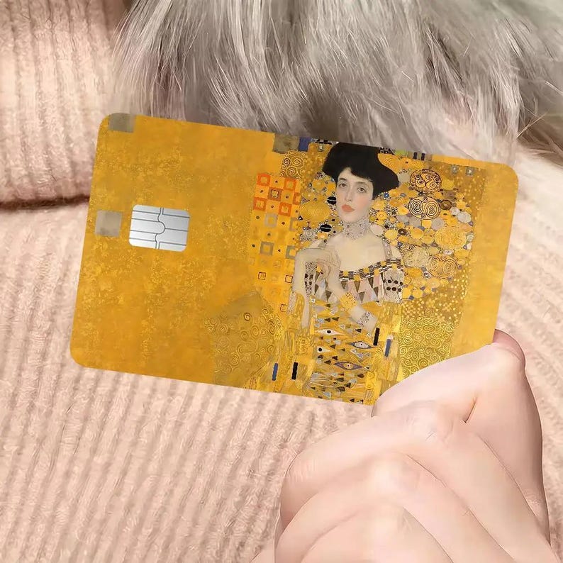 May include: A gold-colored credit card featuring a portrait of a woman in a black hat, with a yellow and gold background. The artwork is reminiscent of Gustav Klimt's style. The card has a chip and is held by a person.