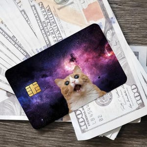 May include: A novelty credit card featuring a galaxy design with a surprised orange and white cat. The card has a gold chip and is resting on a pile of US one hundred dollar bills.