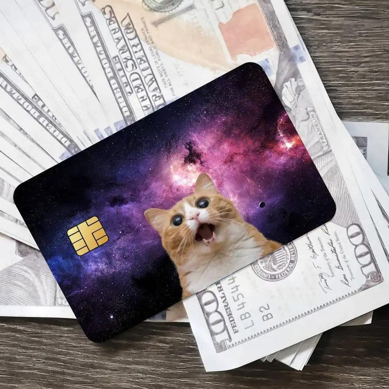 May include: A novelty credit card featuring a galaxy design with a surprised orange and white cat. The card has a gold chip and is resting on a pile of US one hundred dollar bills.
