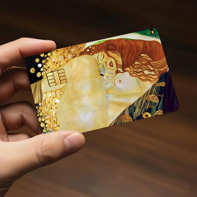 May include: A rectangular card featuring a detail from Gustav Klimt's painting "The Kiss." The artwork depicts a couple embracing, with gold and warm tones dominating the design. The card has a black border and a gold-colored section with a chip.