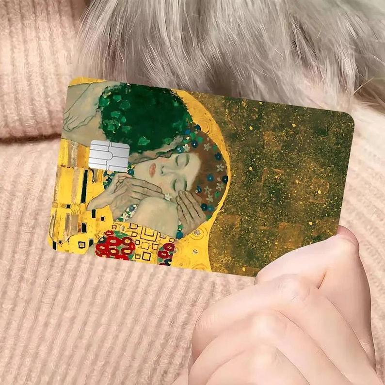 May include: A rectangular credit card featuring a detail from Gustav Klimt's "The Kiss." The design includes gold, green, and red accents, with a depiction of a couple embracing. The card has a white chip and is held by a person.