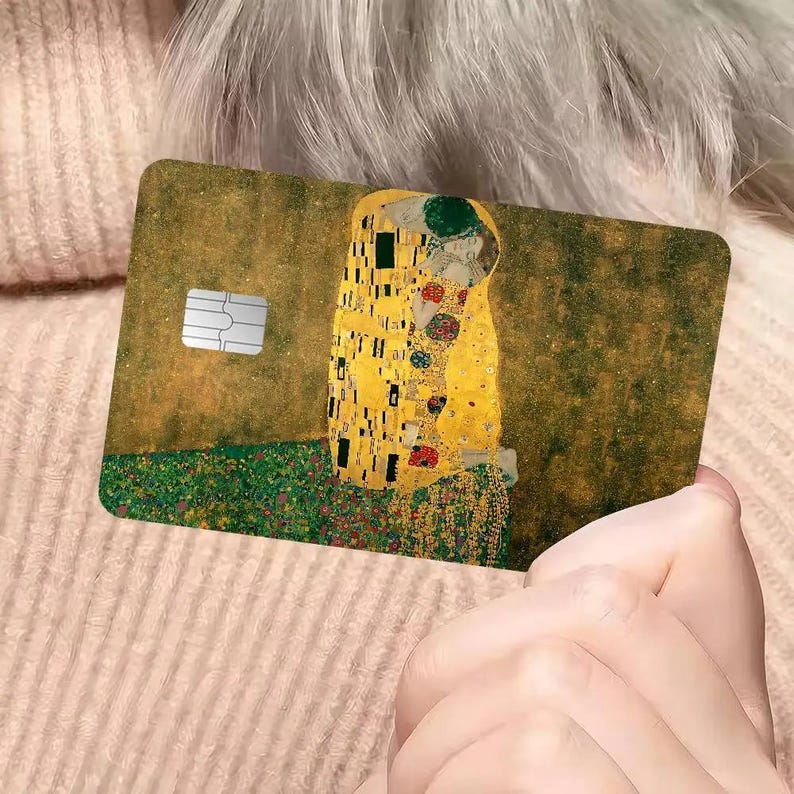 May include: A rectangular card featuring Gustav Klimt's "The Kiss" artwork. The card has a gold and green background with the iconic couple embracing. A silver chip is visible on the left side of the card. The card is held by a person.