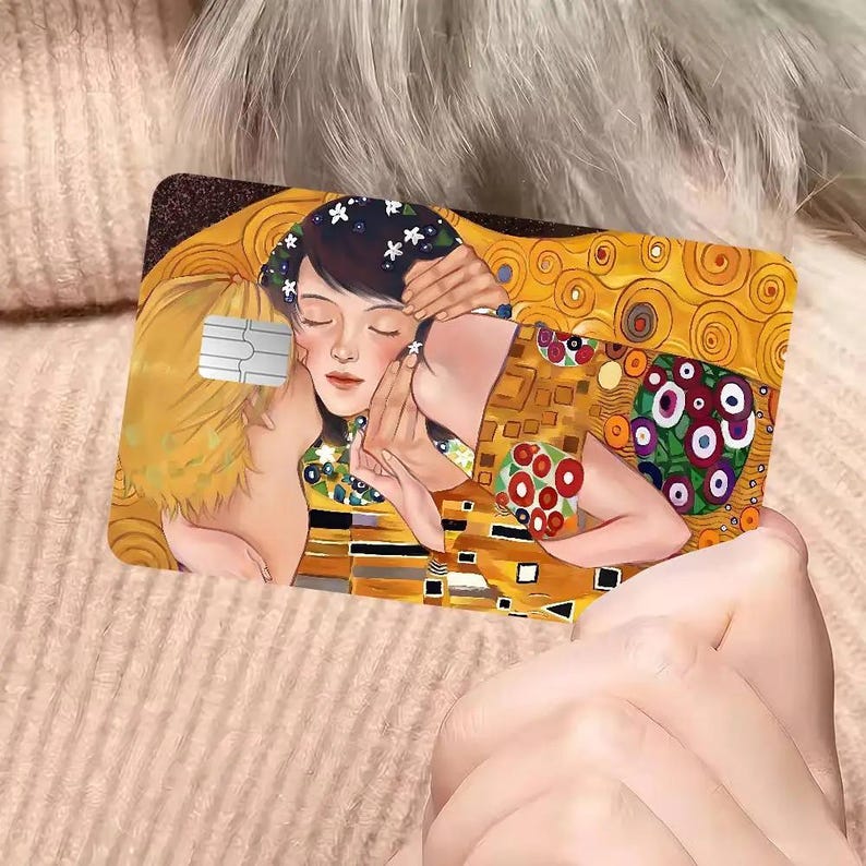 May include: A rectangular card featuring a detail from Gustav Klimt's "The Kiss." The artwork depicts a couple embracing, with the woman adorned with flowers. The card has a gold and brown color scheme, with a white chip on the left side.