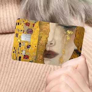 May include: A rectangular card featuring a split design. One side showcases a portion of Gustav Klimt's "The Kiss," with gold and colorful details. The other side depicts a woman's face with gold accents and tears. The card is held in a person's hand.