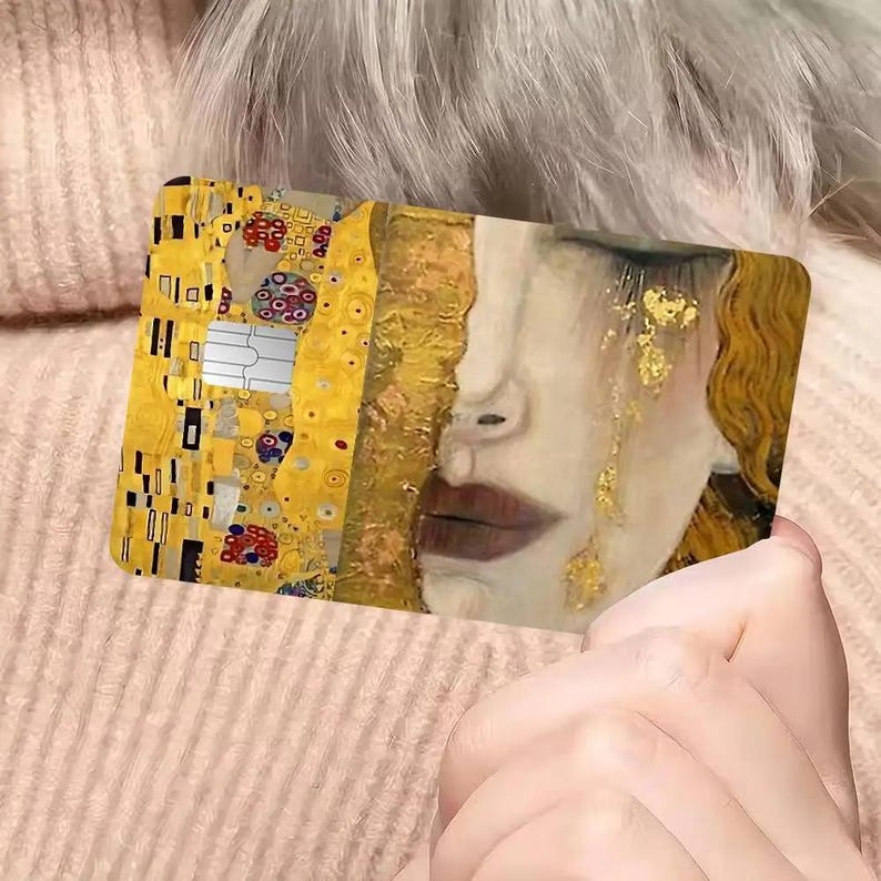 May include: A rectangular card featuring a split design. One side showcases a portion of Gustav Klimt's "The Kiss," with gold and colorful details. The other side depicts a woman's face with gold accents and tears. The card is held in a person's hand.