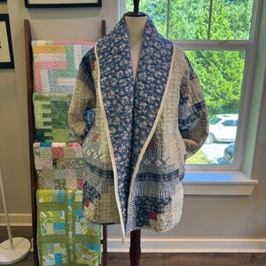 May include: A quilted jacket featuring a patchwork design with various blue, white, and beige fabric patterns. The garment has a wide shawl collar lined with a blue and white floral print, long sleeves, and a light-colored trim along the front edges.