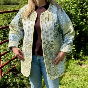 May include: A quilted jacket with a patchwork design in shades of green, blue, and white. The jacket has a round collar and a yellow trim. The person is wearing blue jeans and a brown shirt.