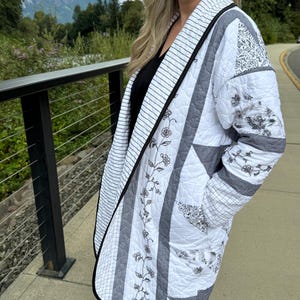 May include: A quilted jacket featuring a patchwork design in white, gray, and black. The garment displays a mix of patterns including thin stripes, floral motifs, and geometric shapes. It has a shawl collar, long sleeves, and a black trim edge.