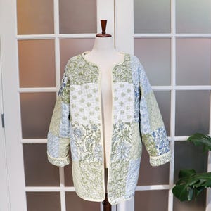 May include: A quilted jacket with a patchwork design. The jacket features a mix of floral and geometric patterns in shades of green, blue, and white. It has a cream-colored trim and is displayed on a mannequin.
