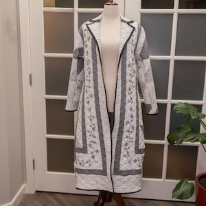May include: A long, quilted coat in white and gray, displayed on a mannequin. The coat features a floral pattern and striped sections, with black trim along the edges. The coat is open, revealing the mannequin's form.