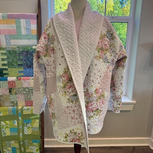 May include: A quilted jacket featuring a patchwork design of floral patterns in shades of pink, green, and white. The garment has a wide shawl collar, long sleeves, and a textured, stitched finish throughout the fabric construction.