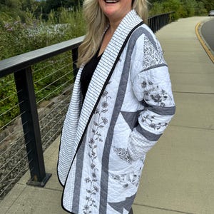 May include: A long-sleeved quilted jacket featuring a patchwork design in white, gray, and black. The garment displays a mix of floral patterns, thin stripes, and geometric star shapes, finished with a black trim along the open front edge.