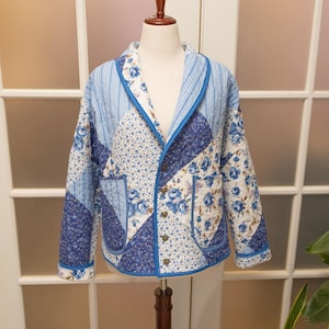 May include: A quilted jacket with a patchwork design. The jacket features a mix of blue floral and striped patterns, with a solid blue trim. It has a button-up front and two pockets. The jacket is displayed on a mannequin.