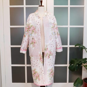 May include: A long, quilted robe with a floral pattern in shades of pink, green, and white. The robe has a relaxed fit, patch pockets, and pink trim along the edges. The robe is displayed on a mannequin.
