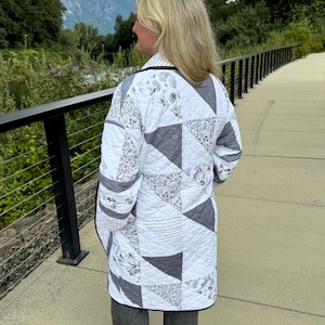 May include: A long-sleeved quilted jacket featuring a patchwork design of white and gray fabric panels. The pattern includes various floral prints, solid gray triangles, and thin black-and-white striped sections, all finished with a black trim.
