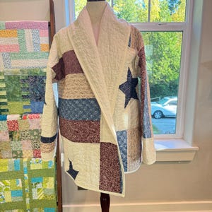 May include: A quilted jacket featuring a patchwork design with cream, navy blue, and burgundy fabric panels. The garment includes a shawl collar, long sleeves, and a prominent navy blue star motif on the front, all finished with detailed stitching.