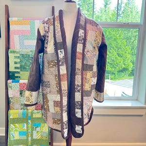 May include: A long-sleeved jacket constructed from a patchwork of various fabrics in shades of brown, cream, and tan. The garment features a dark brown solid-colored trim along the front opening and cuffs, with a quilted texture throughout.