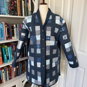 May include: A navy blue quilted jacket with a patchwork design of various blue and white patterns. The jacket has a shawl collar, long sleeves, and a button-up front. The jacket is displayed on a mannequin.