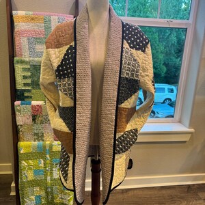 May include: A long-sleeved quilted jacket featuring a patchwork design with cream, tan, and dark blue fabric panels. The jacket has a wide shawl collar with a textured black and white pattern and a black trim along the open front edges.