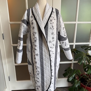 May include: A long-sleeved quilted coat featuring a white and gray color palette. The garment displays a mix of vertical stripes, floral vine patterns, and grid motifs, finished with a black trim along the shawl collar and front opening.