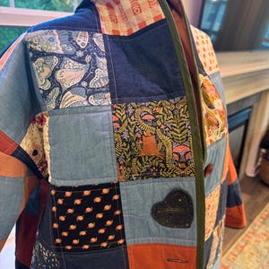 May include: A patchwork jacket constructed from various fabric squares featuring denim, orange, and blue tones. Patterns include mushrooms, snails, floral motifs, and a checkered print. A dark heart-shaped patch is stitched onto the front panel.