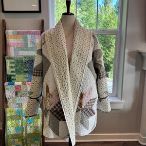 May include: A long-sleeved quilted jacket featuring a patchwork design with various floral and plaid patterns in muted tones of cream, brown, and soft pink. The garment has a wide shawl collar and an open front, constructed from stitched fabric.