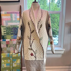 May include: A long-sleeved quilted coat featuring a cream-colored base with a repeating small-scale pattern. The front displays a prominent vine appliqué with brown stems and multi-colored fabric leaves and a single red flower.