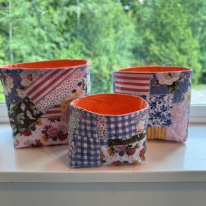 May include: Three fabric storage baskets of varying sizes featuring a patchwork design with floral, striped, and checkered patterns in shades of blue, pink, white, and yellow. Each basket has a bright orange interior lining and visible stitching.