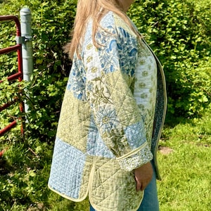 May include: A quilted patchwork jacket in shades of green, blue, and cream. The jacket features floral and geometric patterns, with a light yellow trim. The jacket is worn over blue jeans.