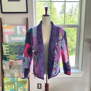 May include: A quilted jacket featuring a patchwork design with various shades of purple, pink, and blue fabric squares. The garment has a shawl collar, long sleeves, and a front-opening construction, displayed on a mannequin torso.