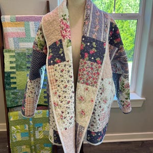 May include: A long-sleeved quilted jacket featuring a patchwork design of various floral and gingham patterns in shades of pink, blue, cream, and green. The garment has a wide shawl collar and an open front, displayed on a mannequin.