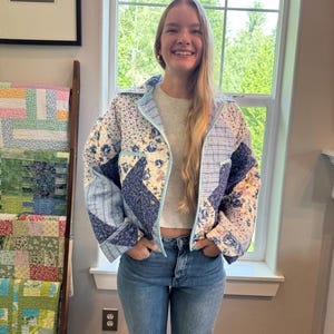 May include: A quilted jacket featuring a patchwork design of blue and white floral patterns mixed with light blue striped fabric. The jacket has a collared neckline, long sleeves, and a front button closure with light blue trim along the edges.